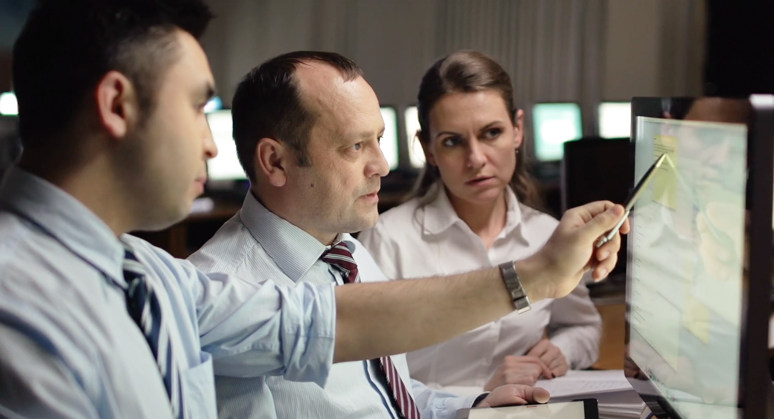 Three business professionals are engaged in a discussion around a computer screen. The person in the center, holding a pen, is pointing at the screen while explaining something. The individual on the left, wearing a tie, is attentively listening, while the person on the right, also wearing business attire, is looking at the screen with a focused expression. The setting appears to be an office environment with dim lighting, and computer monitors are visible in the background.