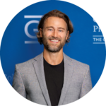A man with wavy hair and a blazer smiling in front of a blue "GW" branded background.
