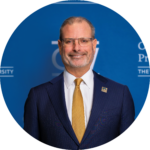 A man in a dark suit and gold tie smiling in front of a blue "GW" branded background.