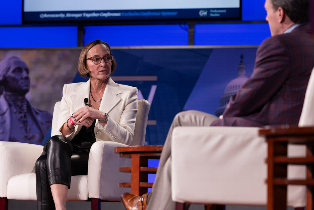 A seated lady, hands crossed, addresses a seated man at the Cybersecurity Stronger Together 2025 conference.