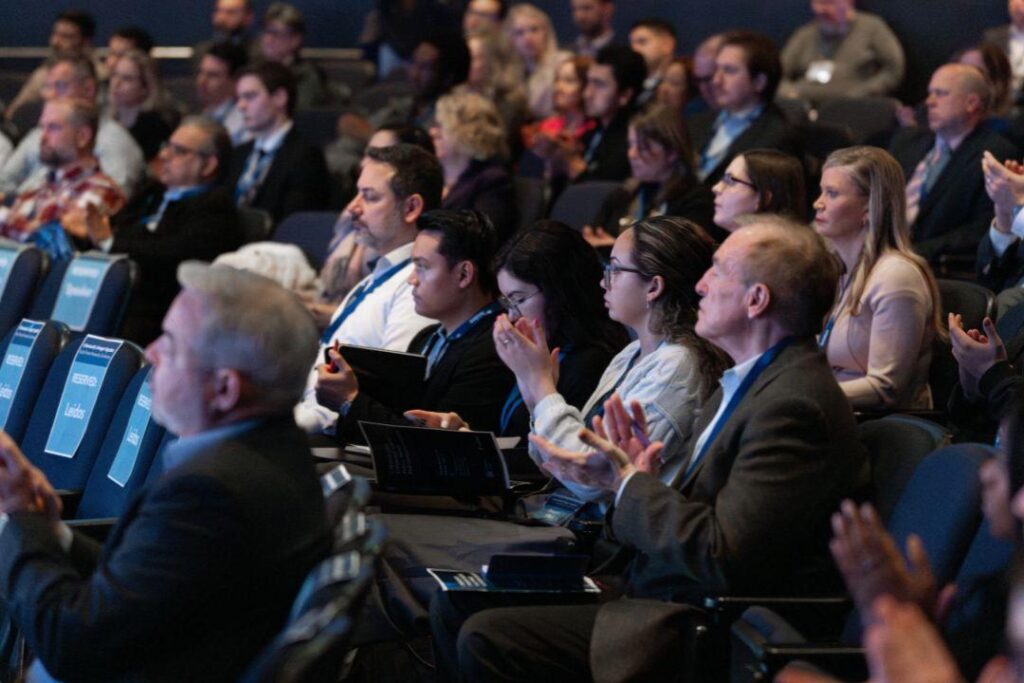 Conference attendees, some of whom are applauding, sit in an auditorium.