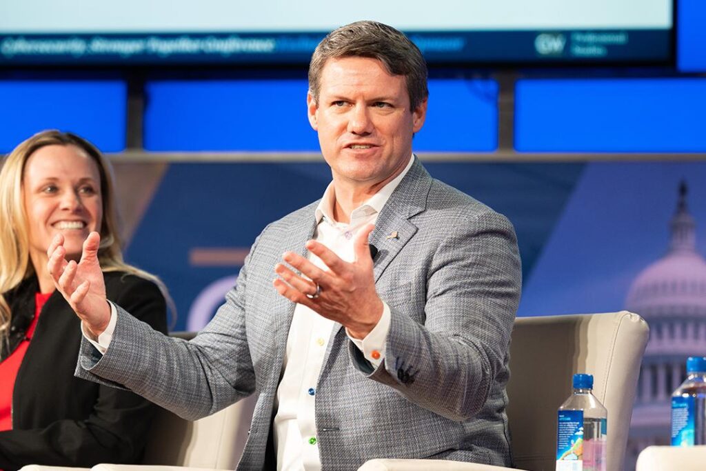 Man in business suit leaning forward in chair and gesticulating with both hands open, palms upturned.